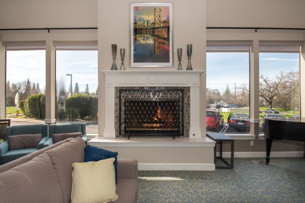 A cozy living room with a modern fireplace, artwork above, and large windows letting in natural light.