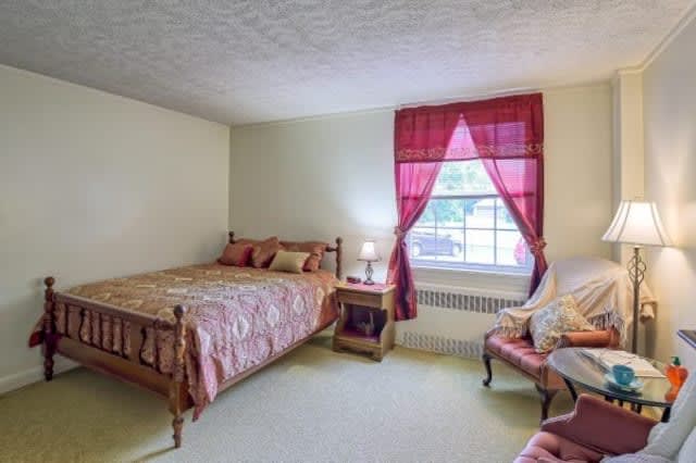 A cozy bedroom with a bed, nightstand, and chair, featuring red curtains.