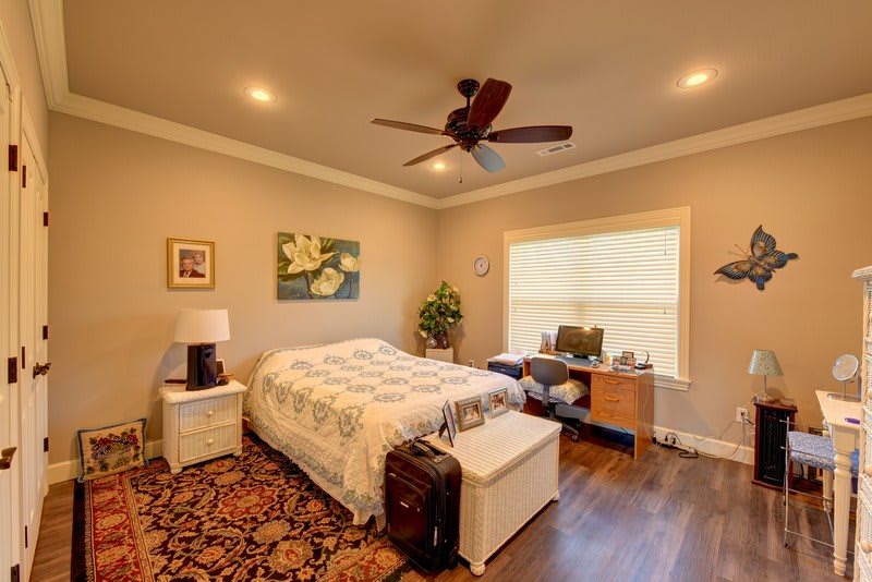 A cozy bedroom with a bed, desk, and ceiling fan, featuring natural lighting and elegant decor.