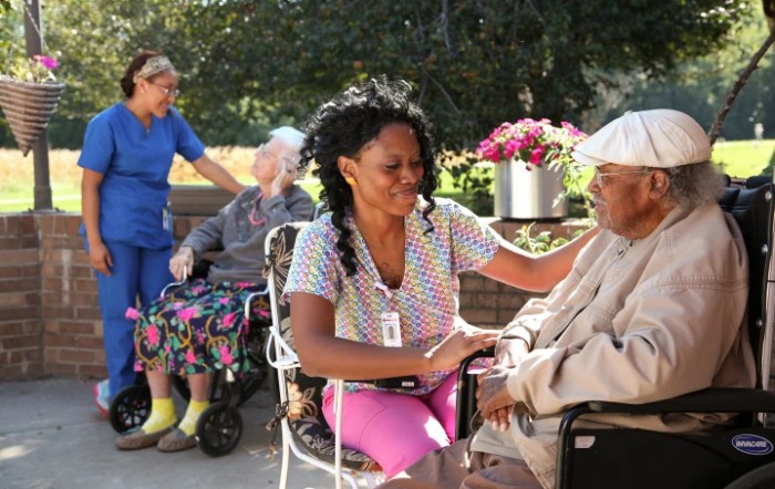 A caregiver interacting warmly with an elderly person in a wheelchair outdoors, alongside others enjoying the day.
