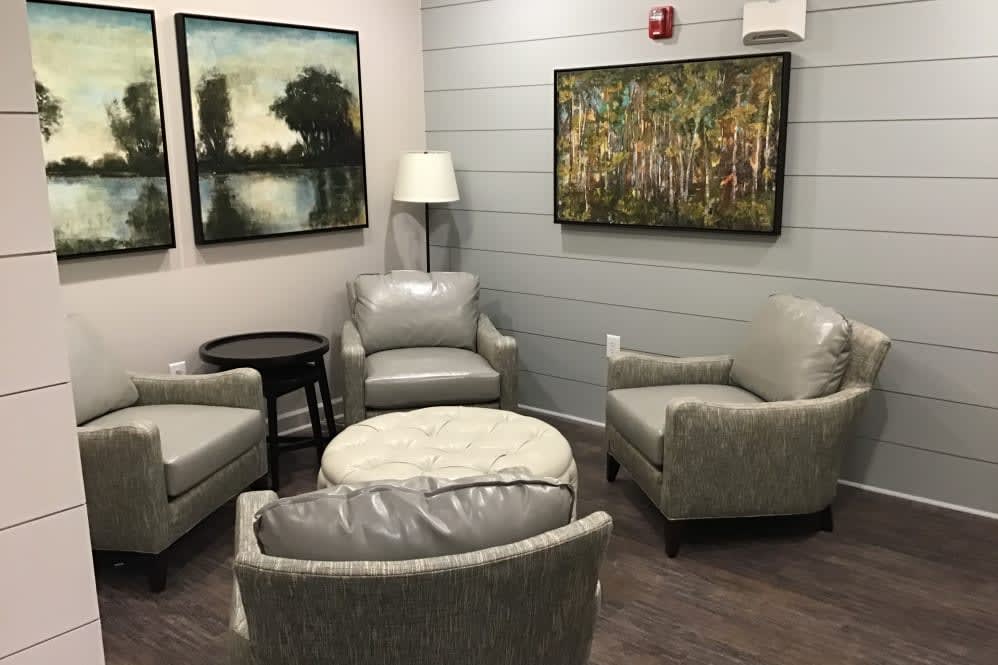 A cozy living room area featuring four gray chairs, a round ottoman, a small side table, a standing lamp, and two landscape paintings on the walls.
