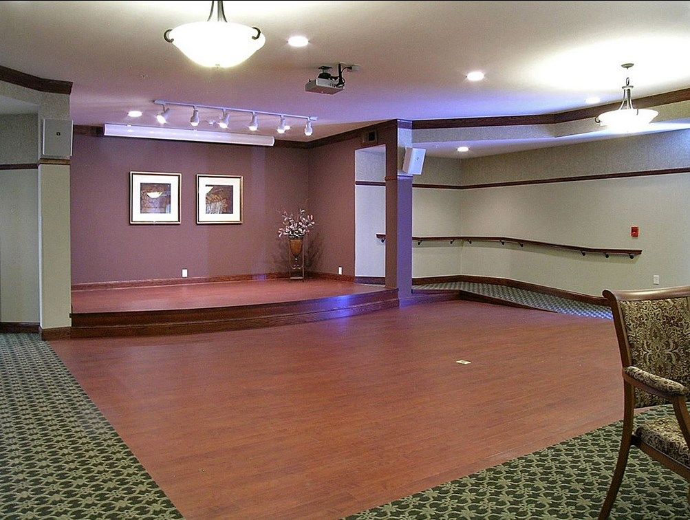 An empty room with wooden flooring and a small stage, featuring ceiling lights and framed artwork.