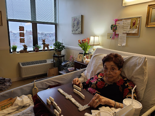 An elderly woman enjoying a game of dominoes in her hospital room, surrounded by plants and personal touches.