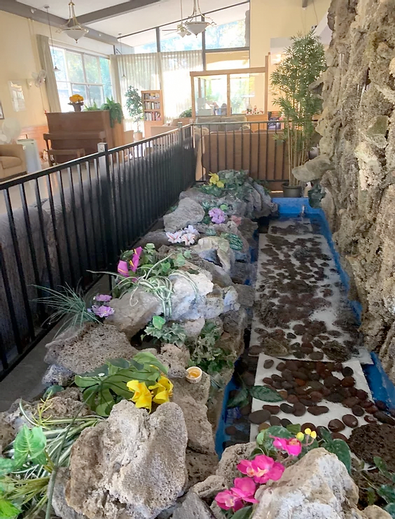 An indoor rock garden with colorful flowers and an artificial pond surrounded by a fence.