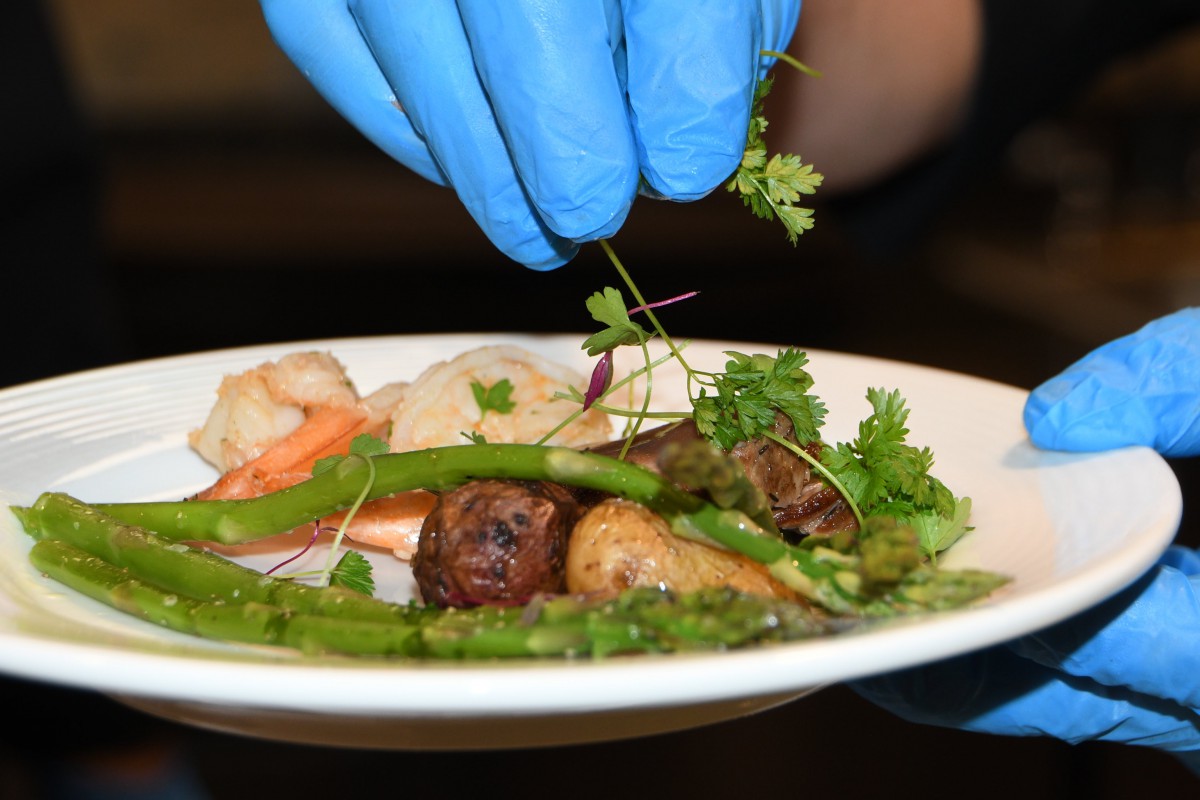 A chef wearing gloves garnishes a dish with fresh herbs and vegetables.