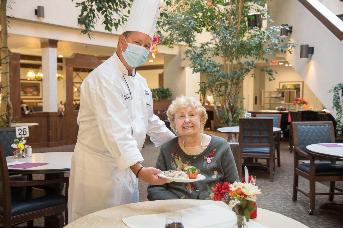 A chef wearing a mask serves a meal to an elderly woman at a restaurant table.