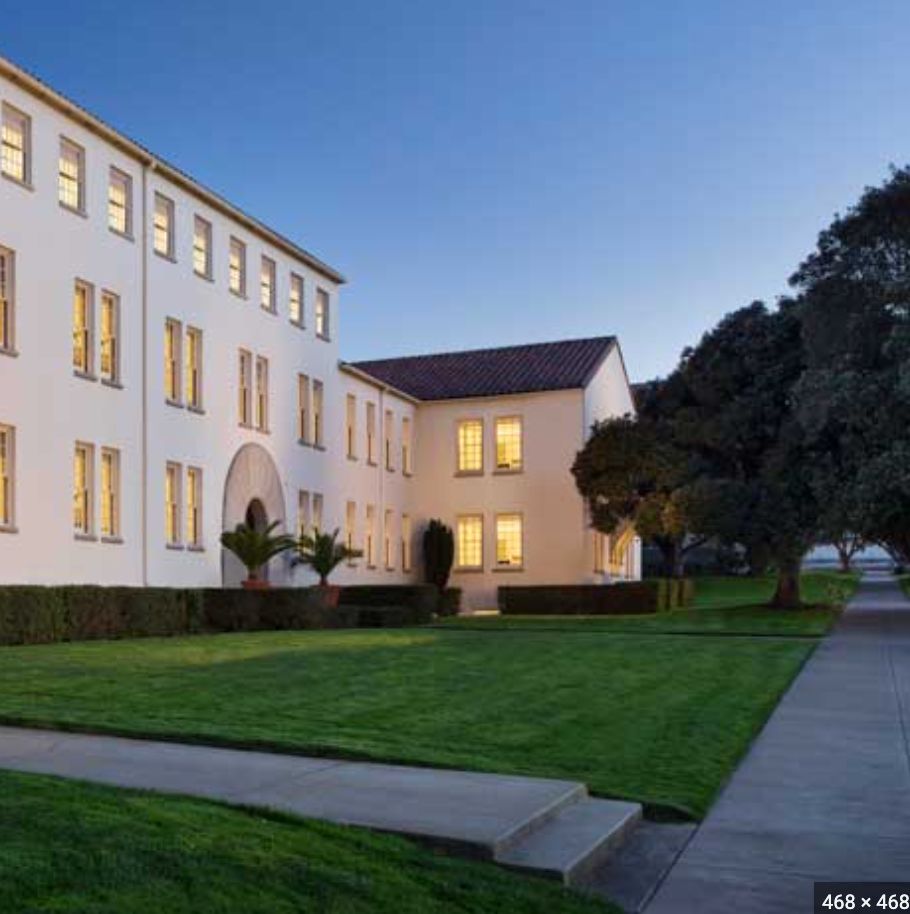 A white building with illuminated windows at dusk, surrounded by a neat lawn and paved pathway.