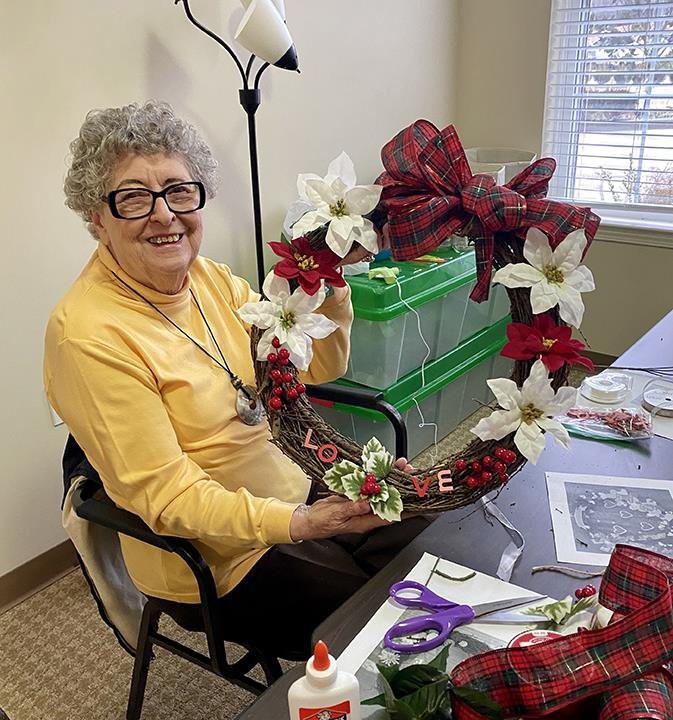 An elderly woman sitting at a table crafting a festive wreath with red and white decorations.