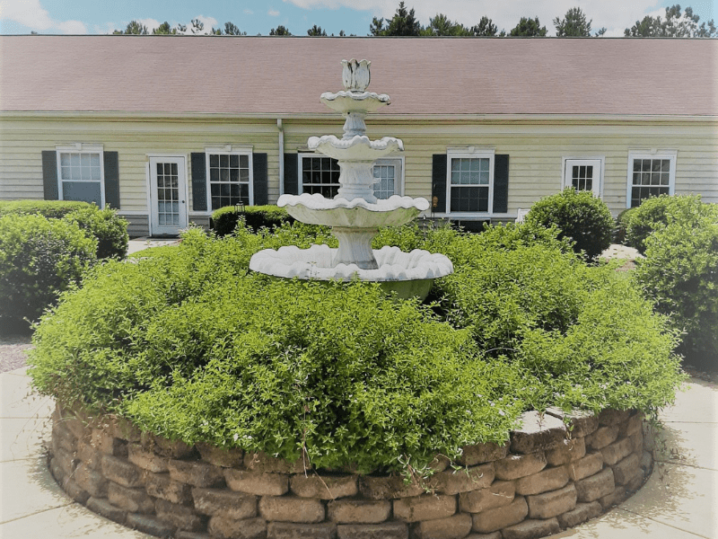 A decorative multi-tiered fountain surrounded by neatly trimmed bushes, in front of a single-story building with a patio.