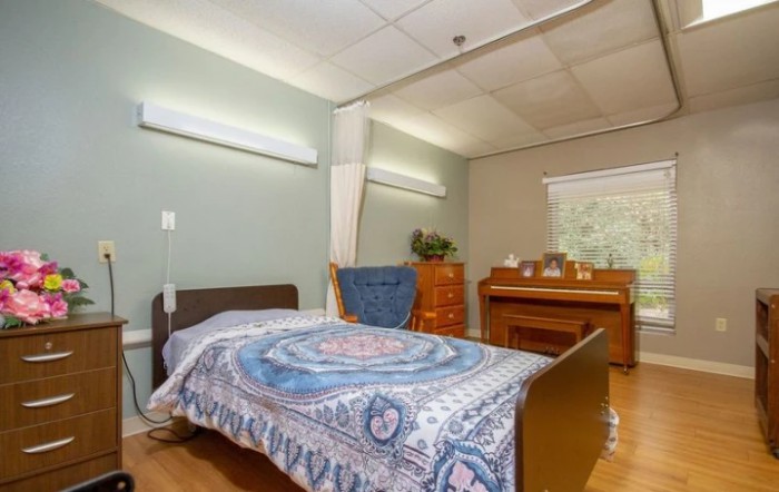 A hospital room with a bed and wooden furniture, featuring a decorative blanket and natural lighting from the window.