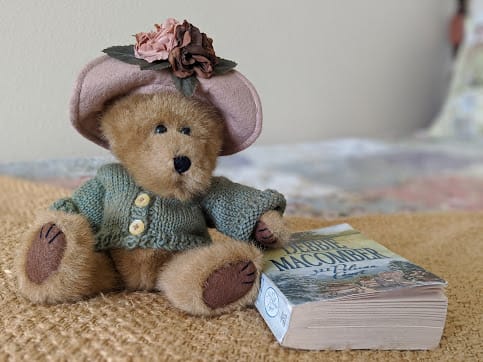 A teddy bear wearing a knitted sweater and hat sits next to a book, creating a cozy atmosphere.
