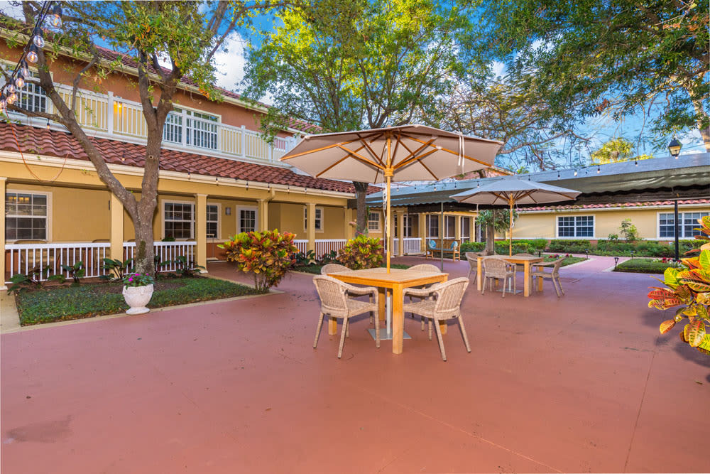 A spacious outdoor patio area featuring tables with umbrellas and chairs, adjacent to a building surrounded by trees on a sunny day.