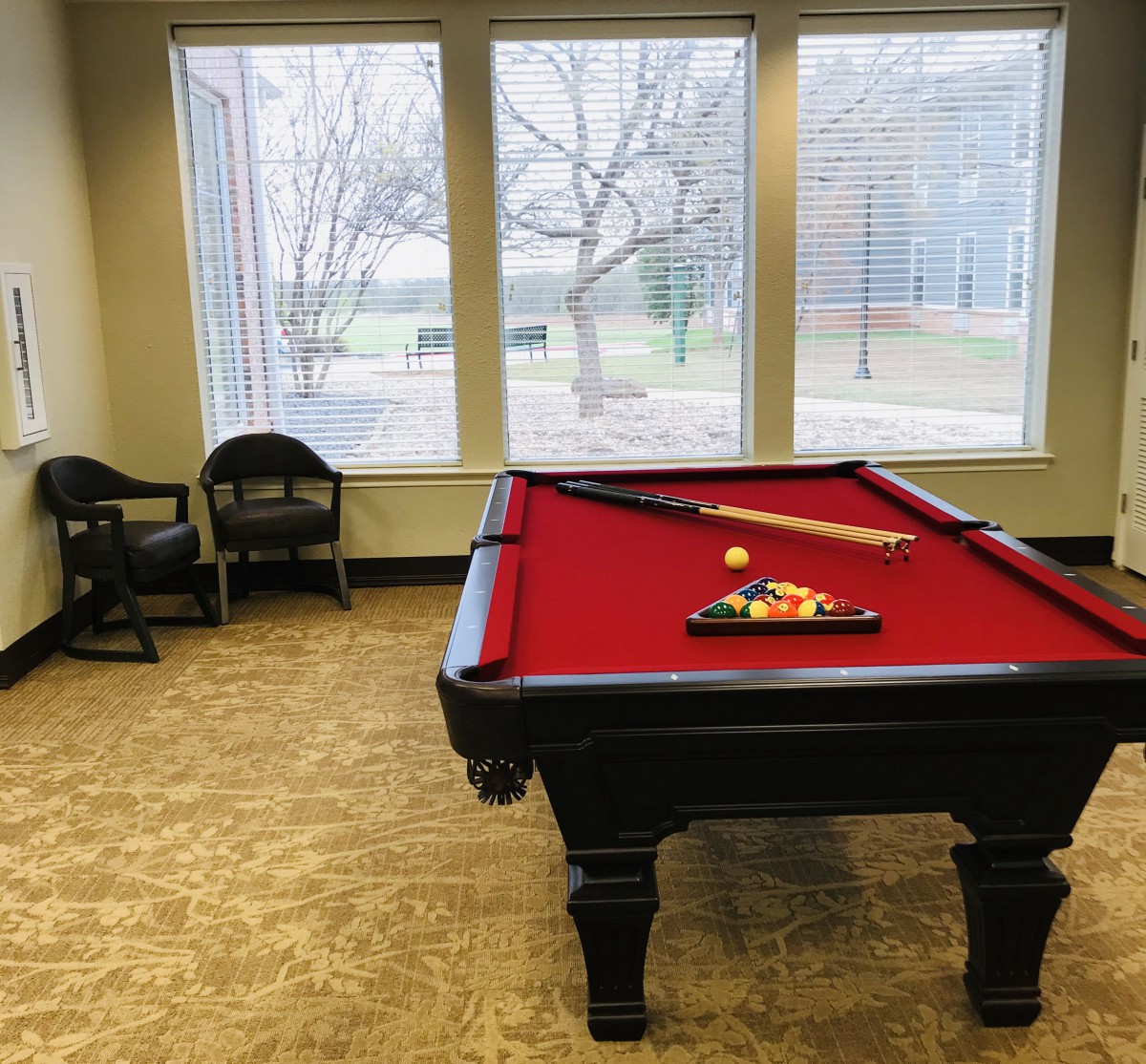A pool table with red felt in a game room featuring large windows and chairs.