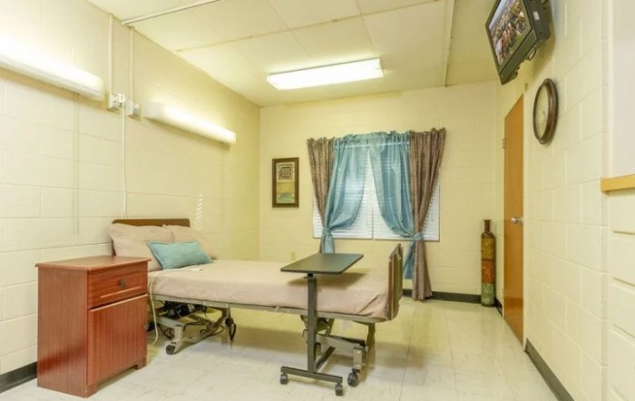 A clean and simple hospital room featuring a single bed, medical equipment, curtains, and a television.