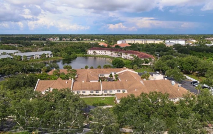 Aerial view of a residential area with buildings surrounded by trees and a lake under a cloudy sky.