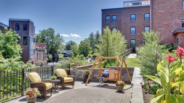 A cozy outdoor patio with a wooden swing and chairs, surrounded by lush greenery and a brick building in the background.