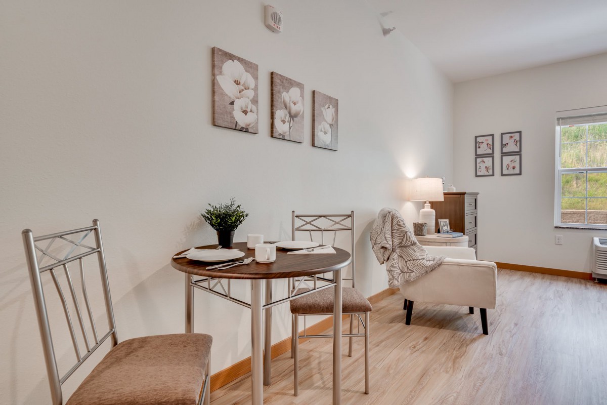 A cozy dining and living area with a small wooden table, two chairs, wall art, and a lamp.