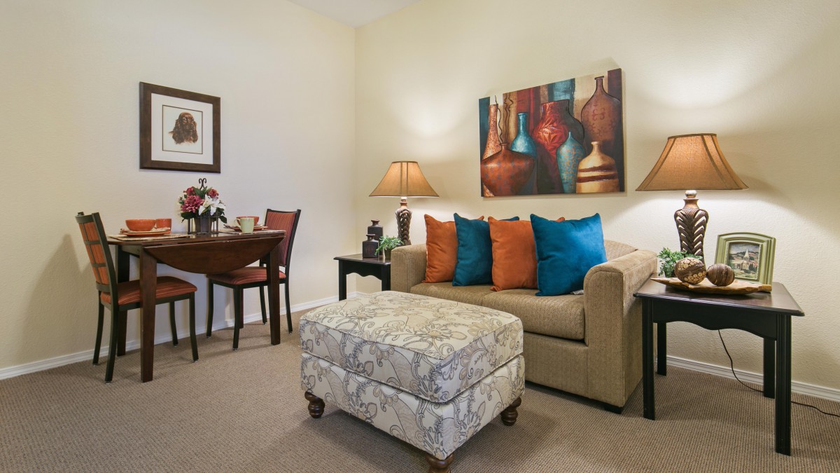 A cozy living room with a sofa adorned with colorful cushions, artwork on the wall, and stylish lamps beside a small dining table.