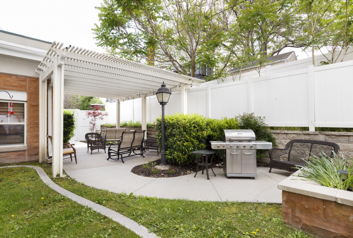 A cozy backyard patio with a pergola, outdoor furniture, and a grill, surrounded by lush greenery and a white fence.