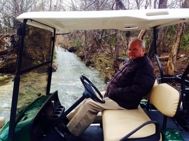 A man sitting in a golf cart near a river with trees surrounding the area.