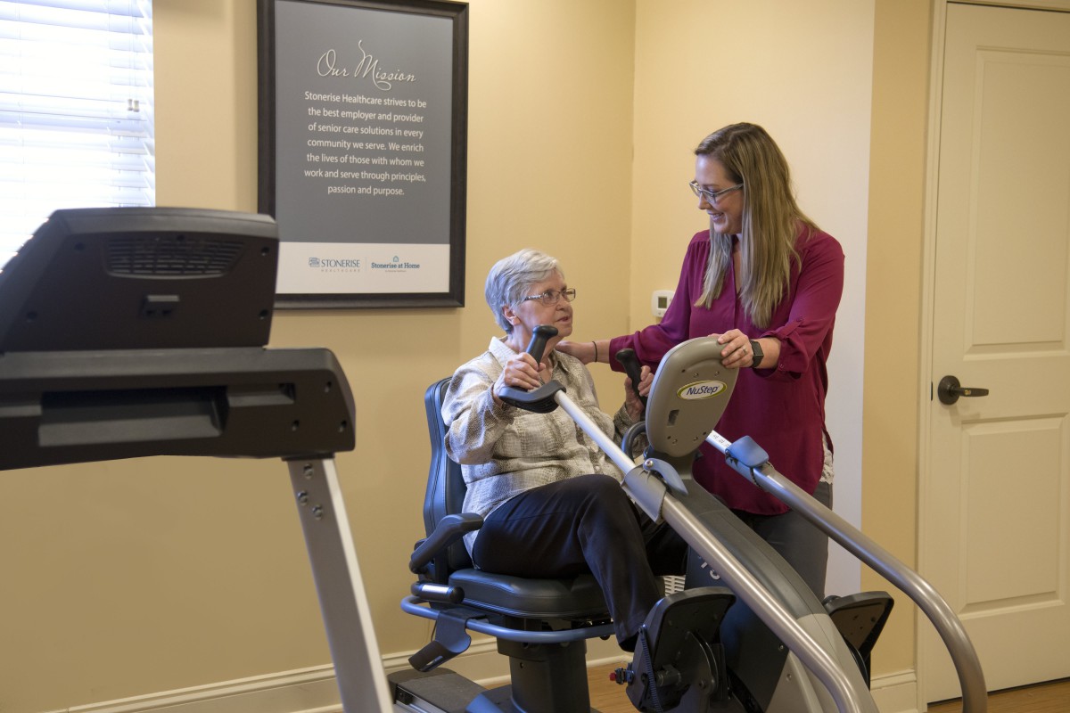 An elderly woman receives assistance from a trainer while using exercise equipment in a gym.