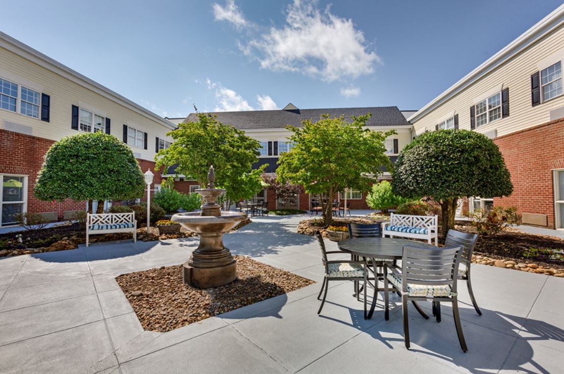A tranquil courtyard featuring a central fountain, surrounded by trees, benches, and tables with chairs.