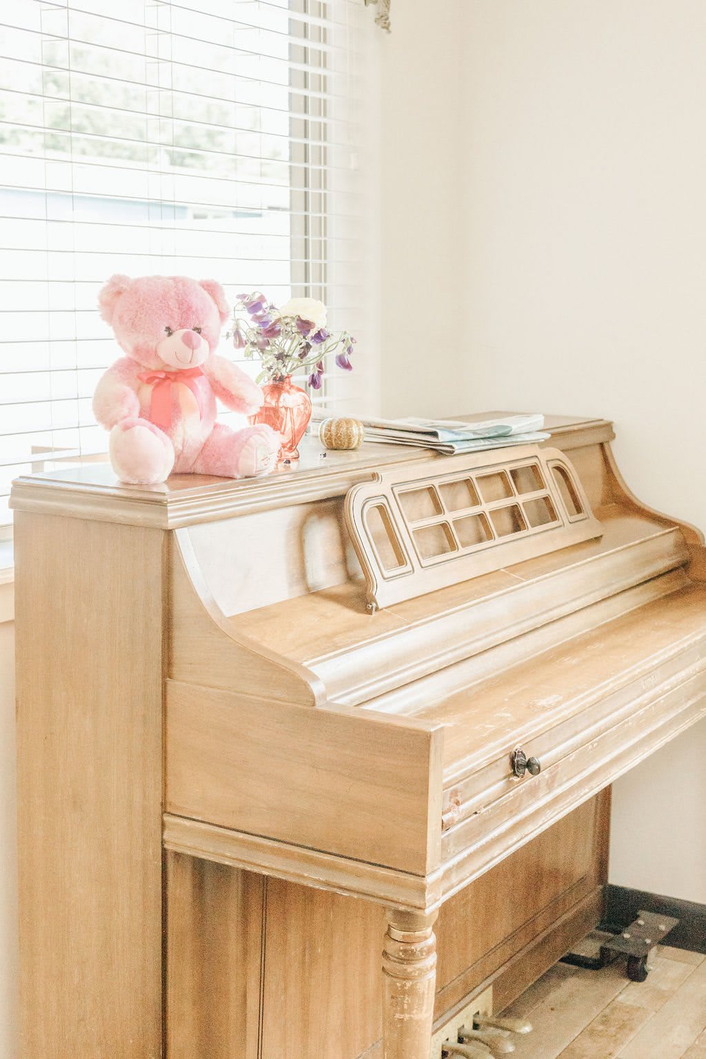A cozy corner with a vintage piano topped by a pink teddy bear and flowers.