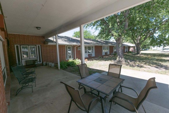 A covered patio area with a table and chairs, adjacent to a brick building and surrounded by greenery.
