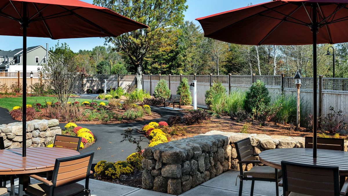A beautifully landscaped outdoor patio with umbrellas, chairs, and a stone wall surrounded by trees.