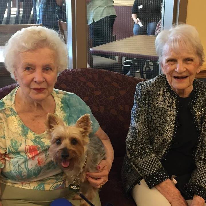 Two elderly women sitting indoors with a small dog, smiling at the camera.