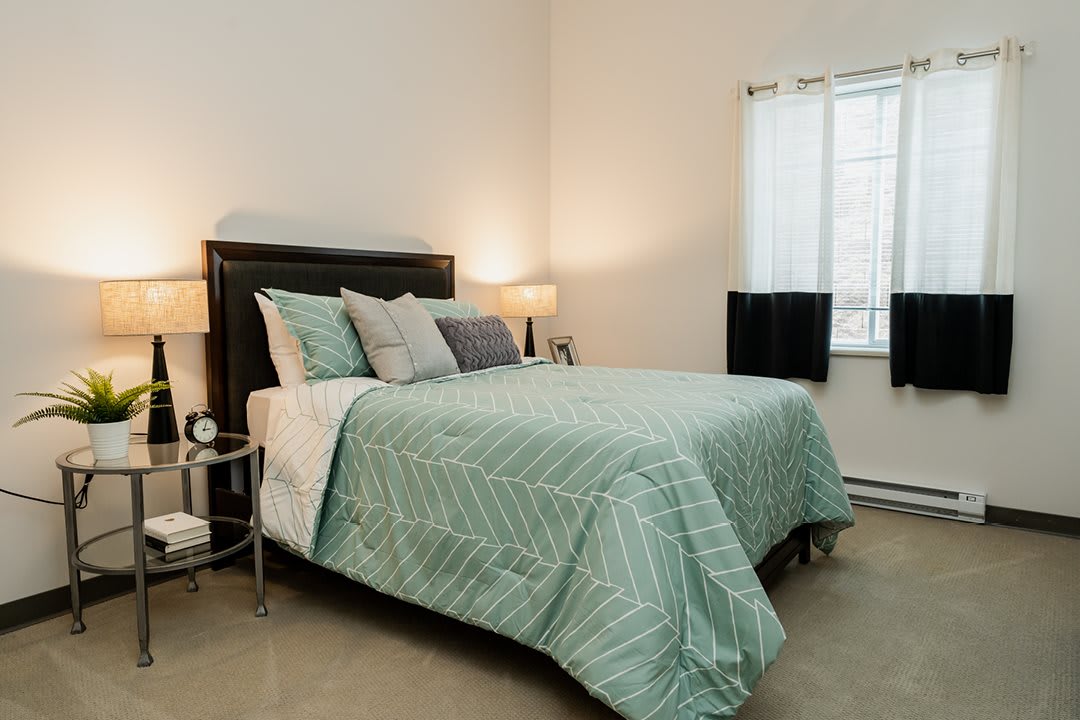 A cozy bedroom with a neatly made bed, featuring a green comforter and several pillows. The room is softly lit by lamps on either side of the bed.