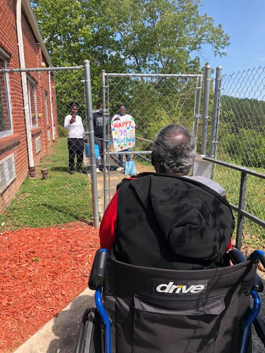 A person in a wheelchair enjoys the outdoors while being assisted by caregivers near a fenced area.