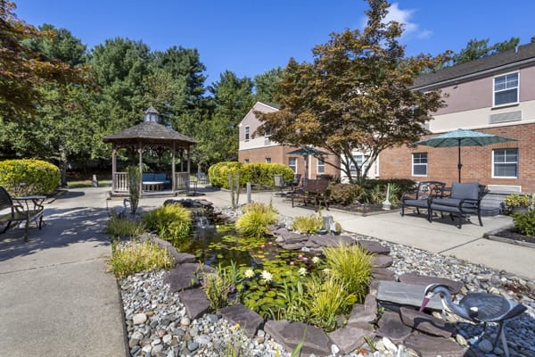 A beautifully landscaped garden featuring a gazebo, a pond surrounded by rocks and greenery, and outdoor seating on a sunny day.