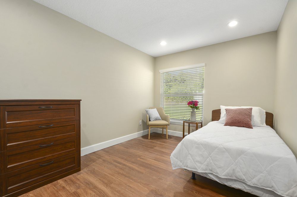 A minimalist bedroom with a comfortable bed, wooden dresser, and large window allowing natural light.