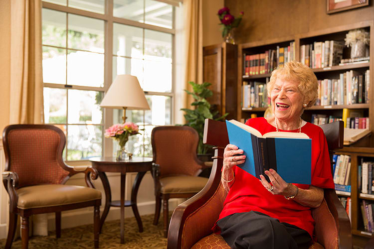 An elderly woman sits in a cozy living room, smiling while reading a book.