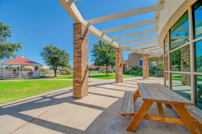 A sunny patio with a pergola and picnic table, surrounded by a green lawn.
