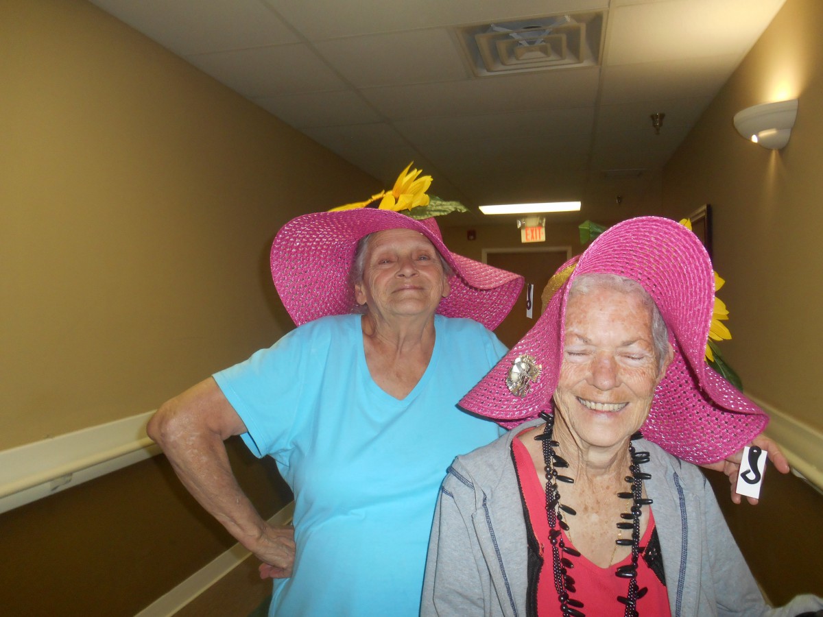 Two elderly women wearing pink hats with flowers are smiling indoors.