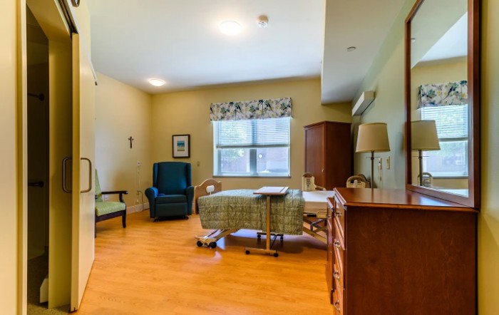 A well-furnished nursing home room with a bed, chair, and window.