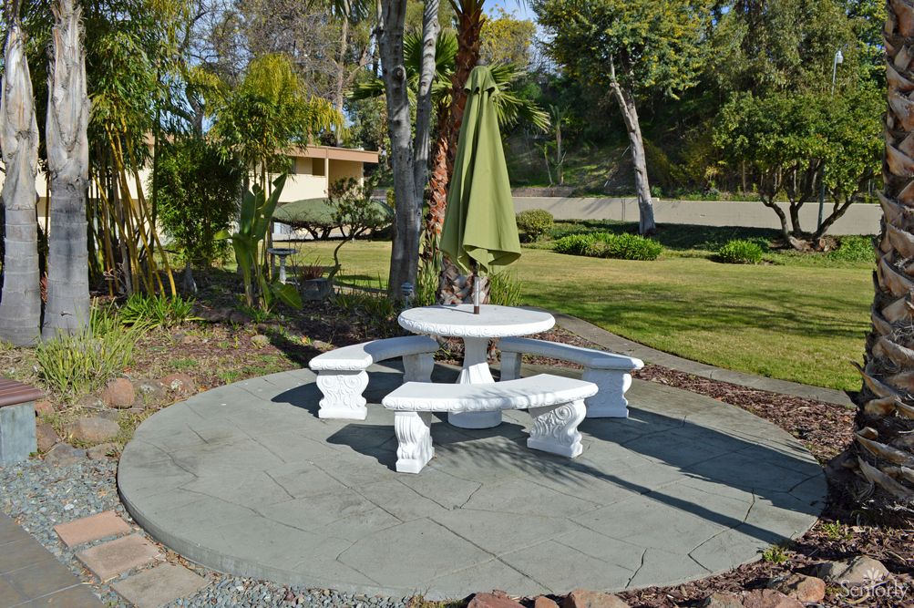 A scenic garden area featuring a white stone table with benches and a green umbrella, surrounded by trees and lush greenery.