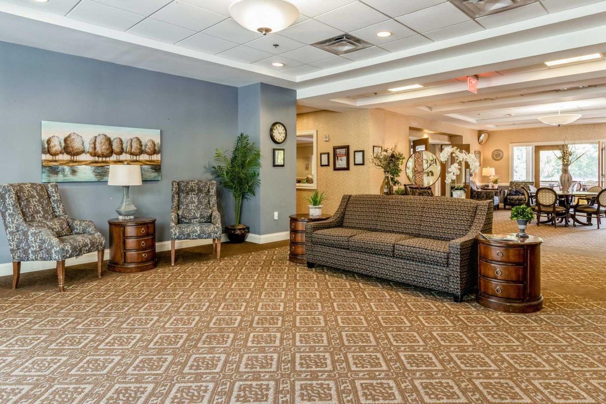A cozy lobby area featuring a patterned carpet, sofa, chairs, wall art, and indoor plants.