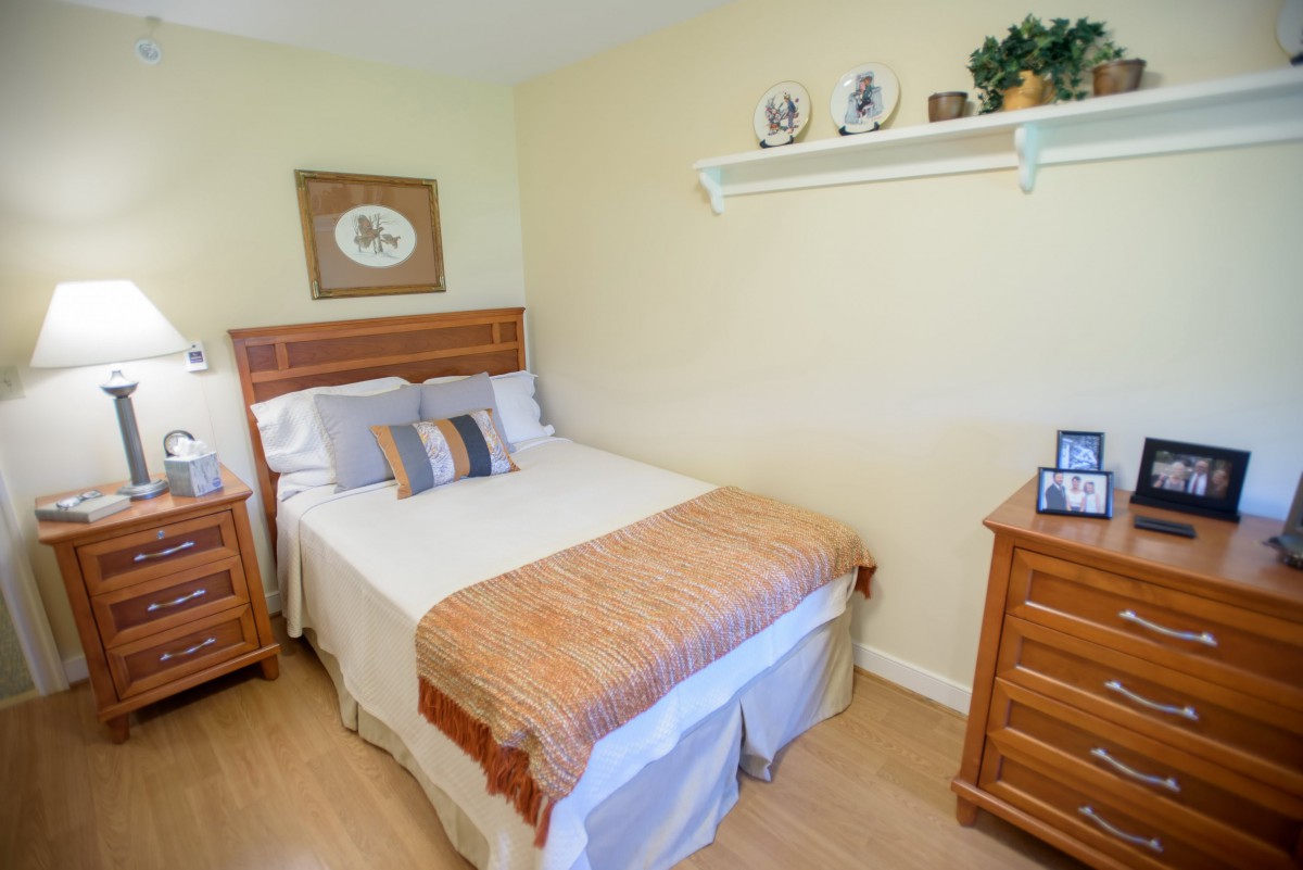 A cozy bedroom with a neatly made bed, wooden furniture, and decorative accents on a shelf.