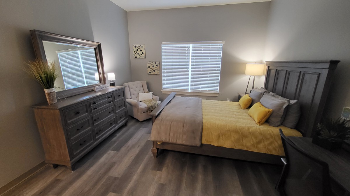A modern bedroom with a bed, dresser, and armchair, featuring yellow accents and natural lighting.