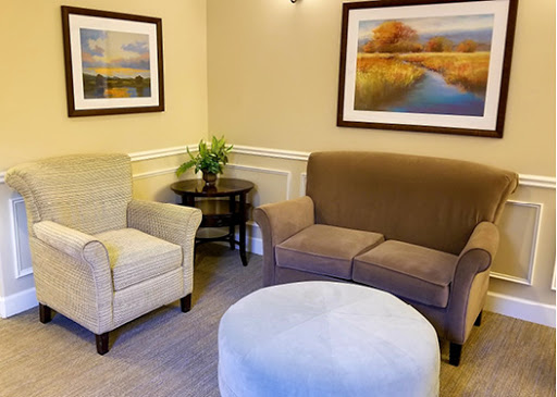 A cozy seating area featuring a light-colored armchair, a brown sofa, an ottoman, and decorative artwork.