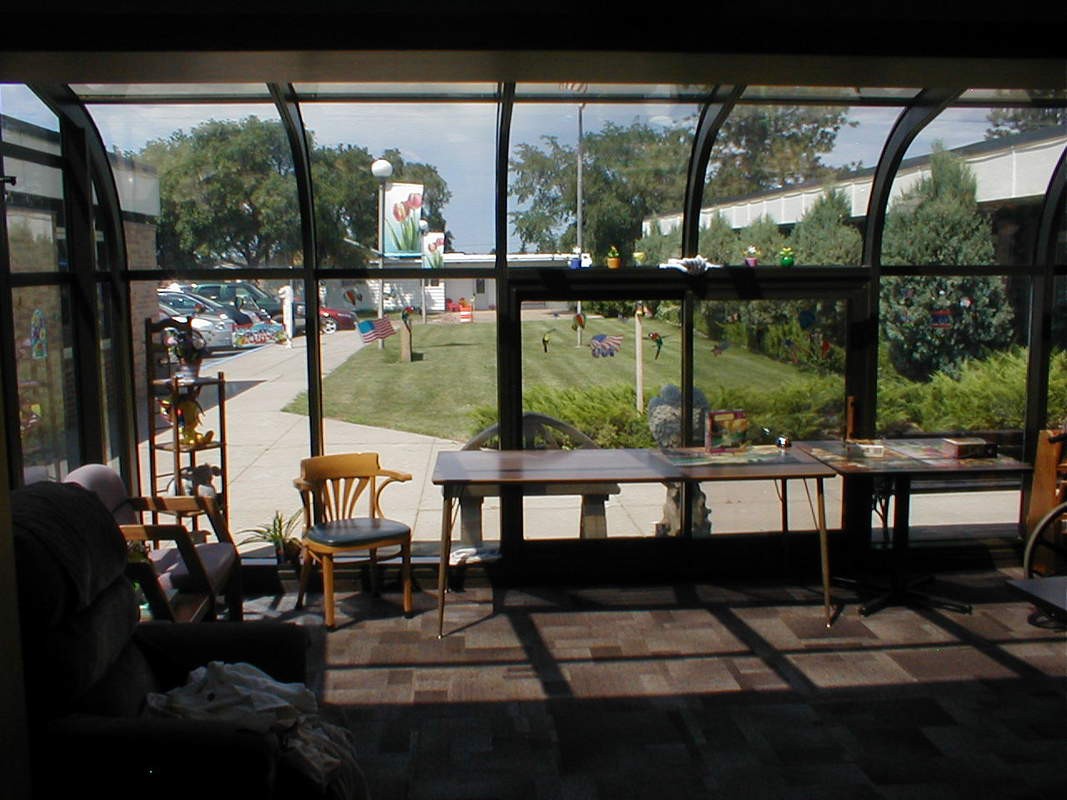 A brightly lit indoor space with large windows overlooking a garden, featuring tables and chairs arranged in the sunlight.