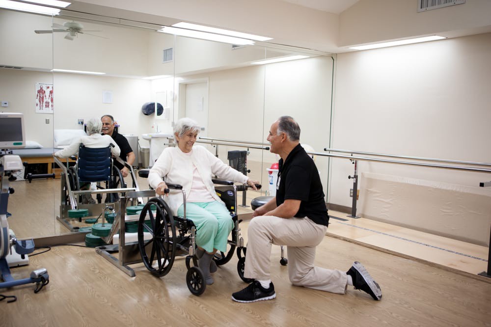 A physical therapist kneels beside an elderly person in a wheelchair, providing support and care in a rehabilitation setting.
