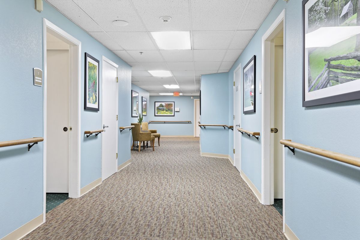 A well-lit hallway with blue walls and carpeted floor, adorned with paintings and featuring several doors on each side.