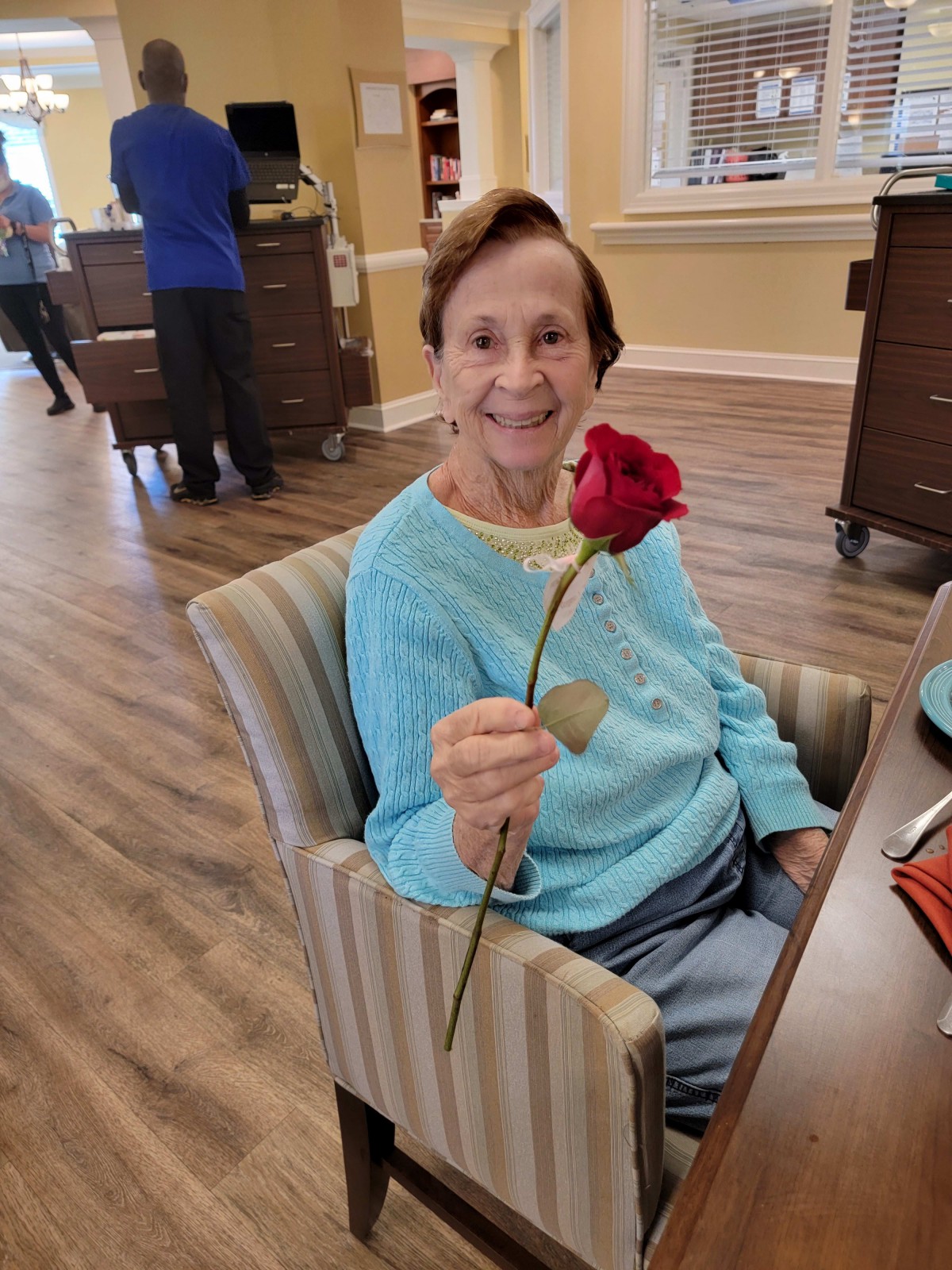 An elderly woman sitting in a chair indoors, holding a red flower and smiling.