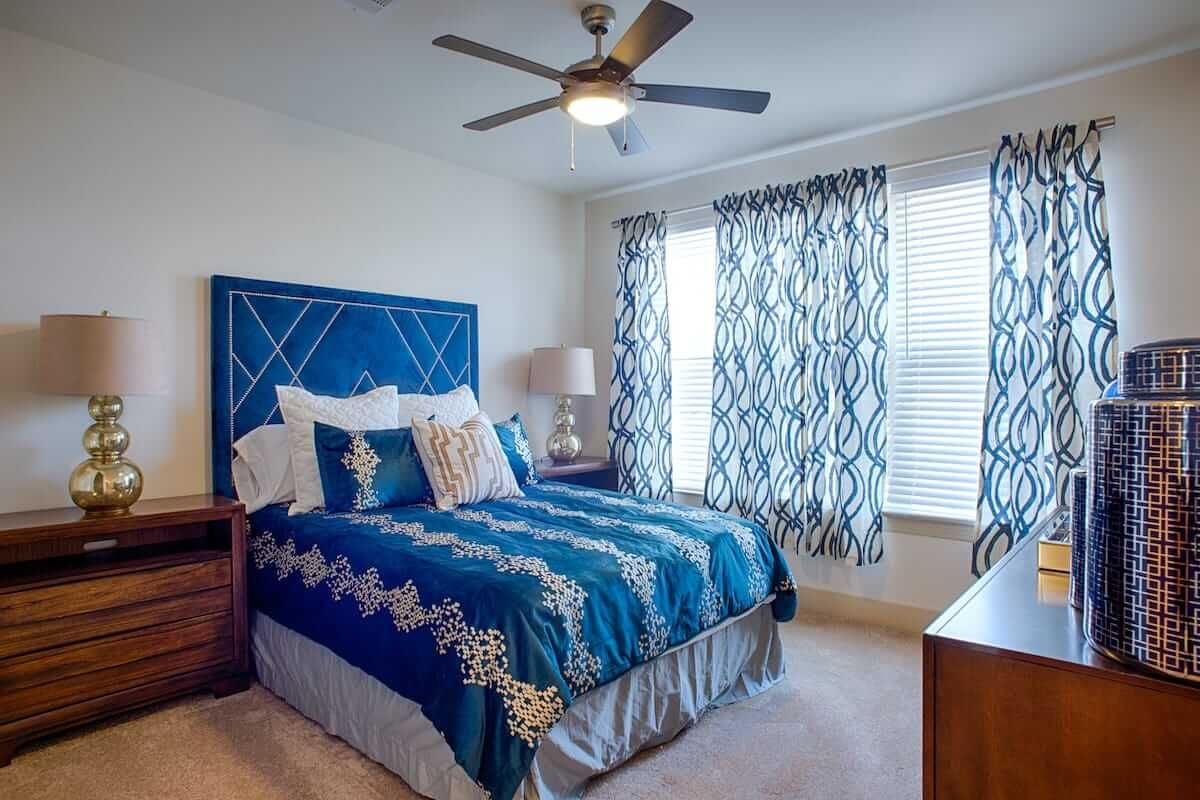 A cozy bedroom featuring blue bedding and patterned curtains, with a ceiling fan above.