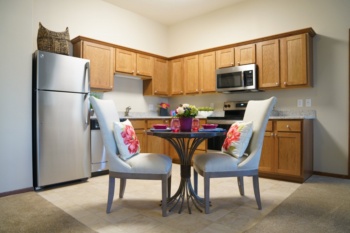 A cozy kitchen with a small dining area featuring a round table and two cushioned chairs, surrounded by wooden cabinets and stainless steel appliances.
