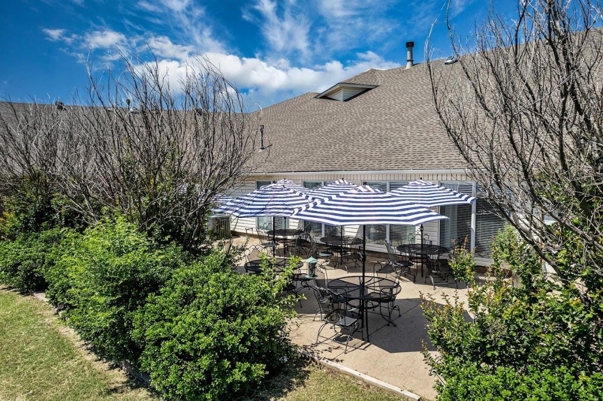 A sunny patio area with striped umbrellas, tables, and chairs surrounded by greenery.